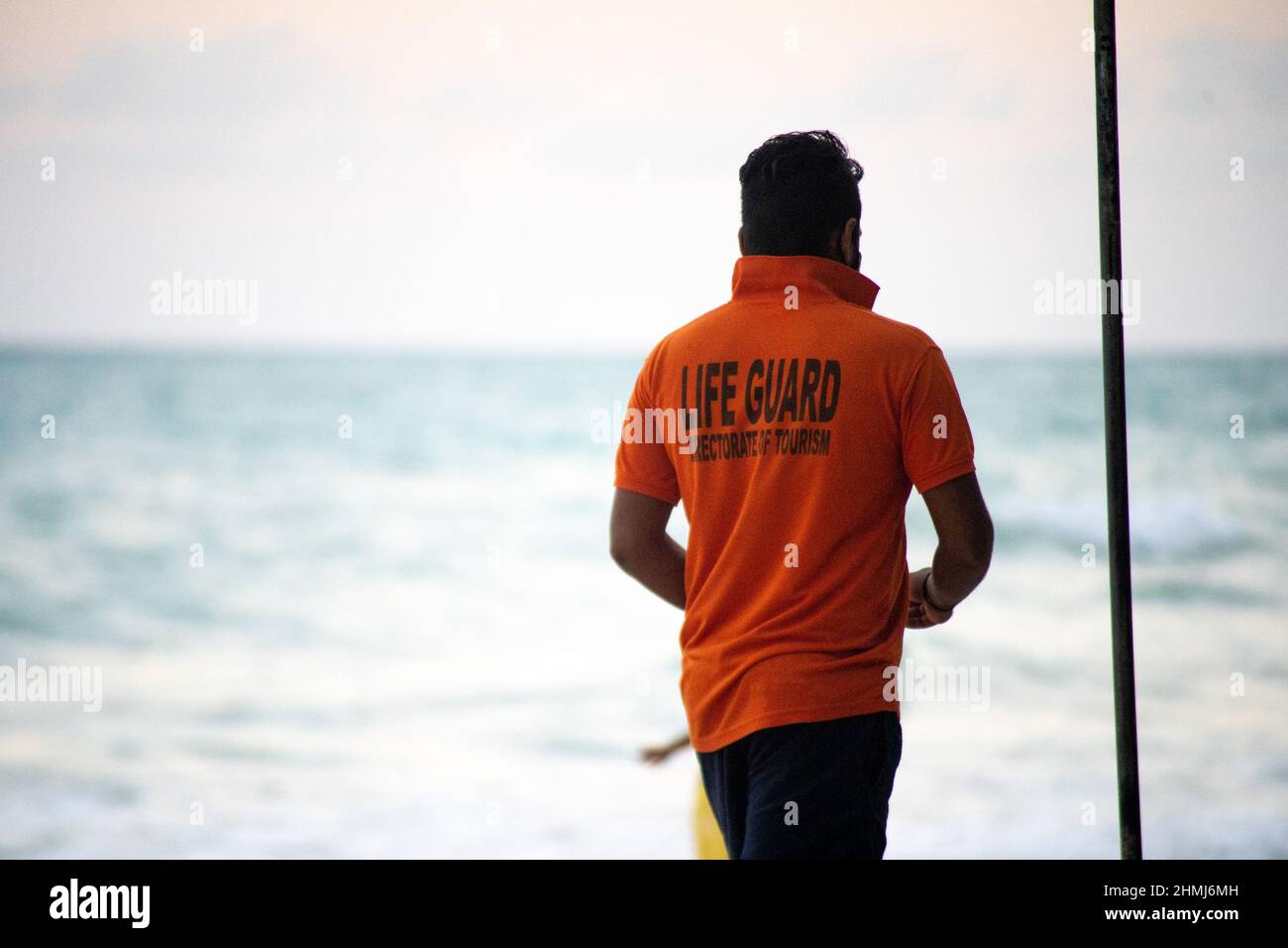 Rettungsschwimmer in orangefarbenem Hemd mit Flagge am Strand entlang mit Touristen im Hintergrund bei havelock swaraj dweep andaman nicobar Islands india Stockfoto