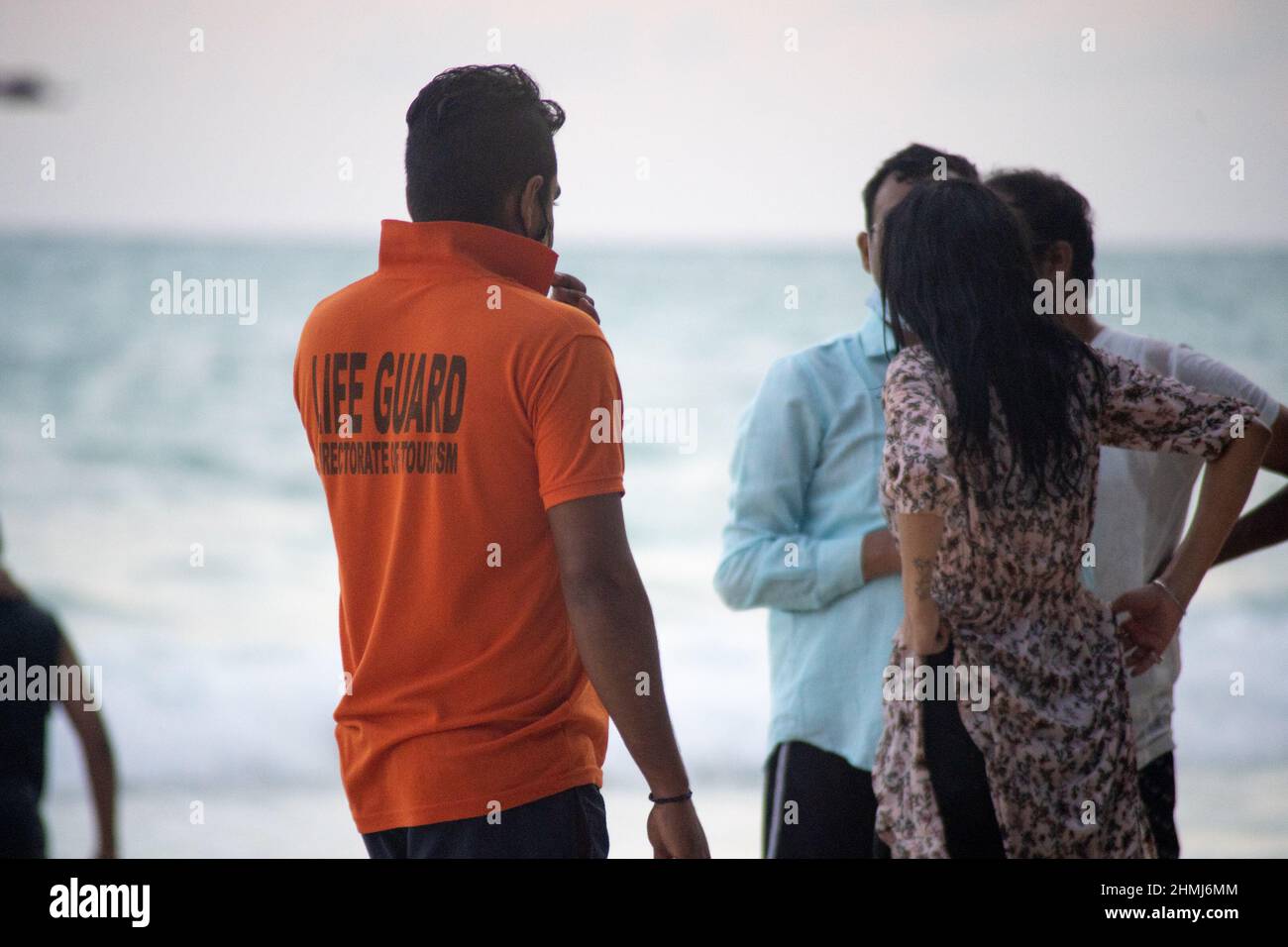 Rettungsschwimmer in orangefarbenem Hemd mit Flagge am Strand entlang mit Touristen im Hintergrund bei havelock swaraj dweep andaman nicobar Islands india Stockfoto