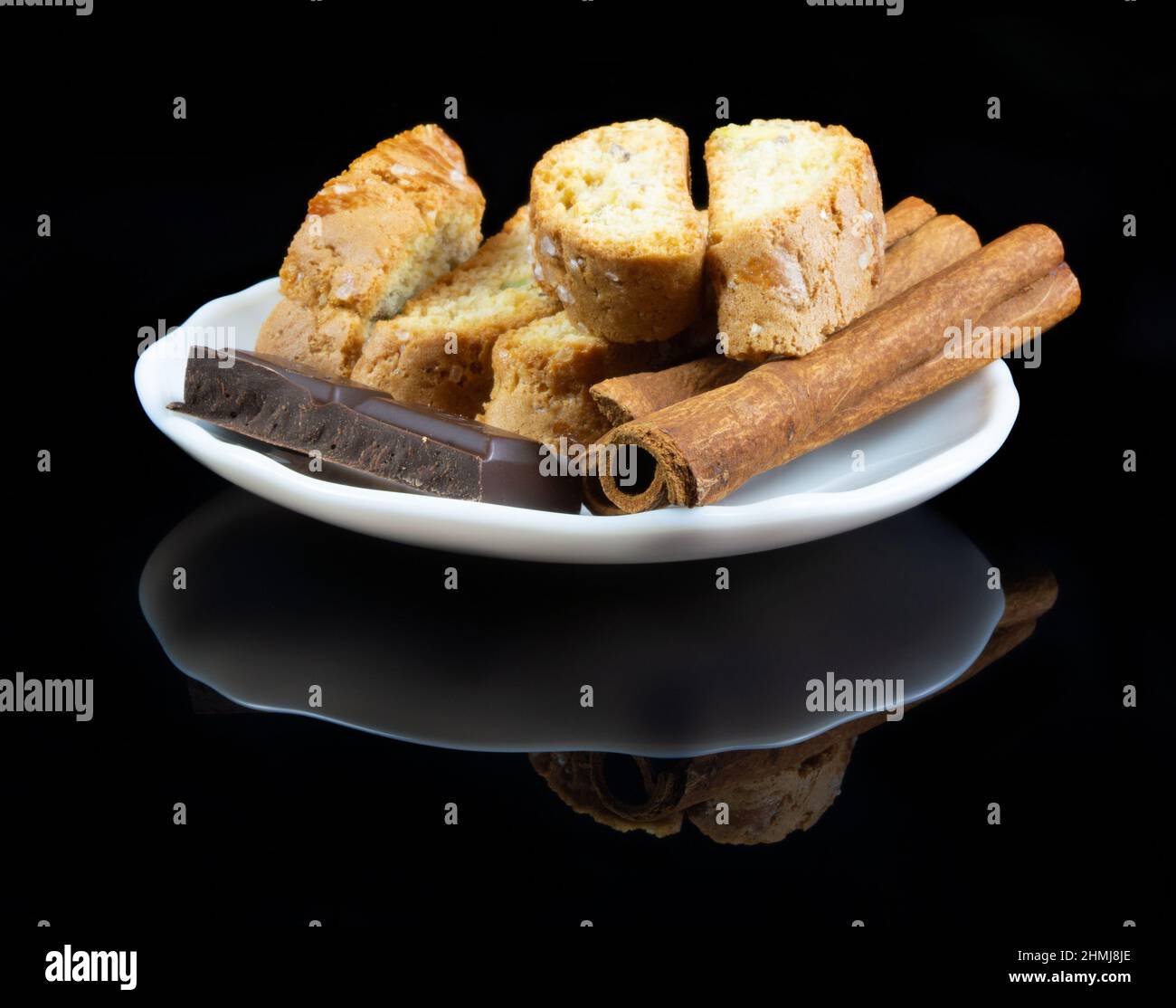 Cinnanon-Sticks, Schokolade und Bisquits auf einem weißen Teller auf schwarzem reflektierendem Hintergrund Stockfoto