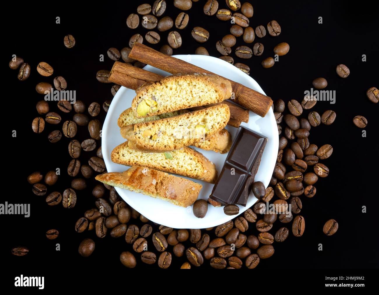 Cinnanon-Sticks, Schokolade und Bisquits auf einem weißen Teller, umgeben von Kaffeebohnen auf schwarzem, reflektierendem Hintergrund Stockfoto