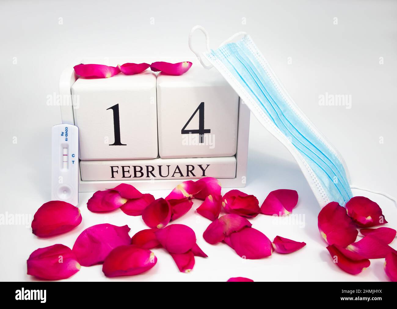 Februar 14 weißer Holzkalender auf weißem Hintergrund mit roten Rosenblättern. Neben ihm befindet sich eine medizinische Maske und ein positiver Covidtest Stockfoto