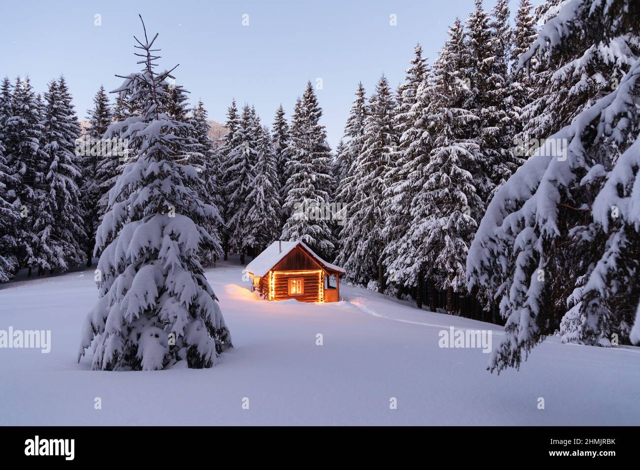 Abends leuchten die Lampen das Haus auf. Winterlandschaft. Holzhütte auf dem mit Schnee bedeckten Rasen. Mystische Nacht. Heiraten Sie Weihnachten und Neu Stockfoto