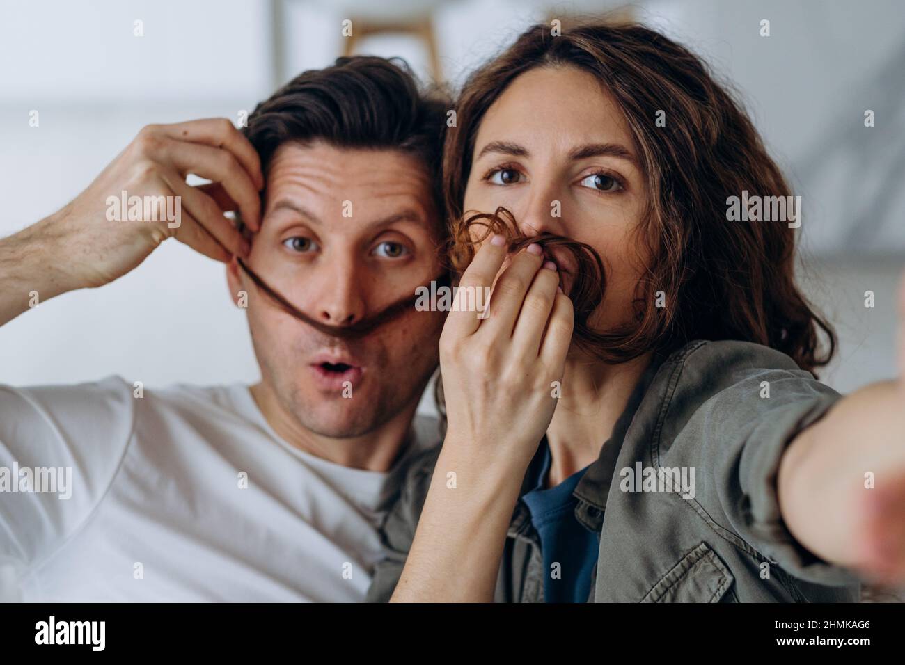 Fröhliches Brautpaar spielt und posiert Schnurrbärte mit lockigen Brünetten-Haaren und küsst lächelnd für Selfies per Smartphone-Nahaufnahme Stockfoto