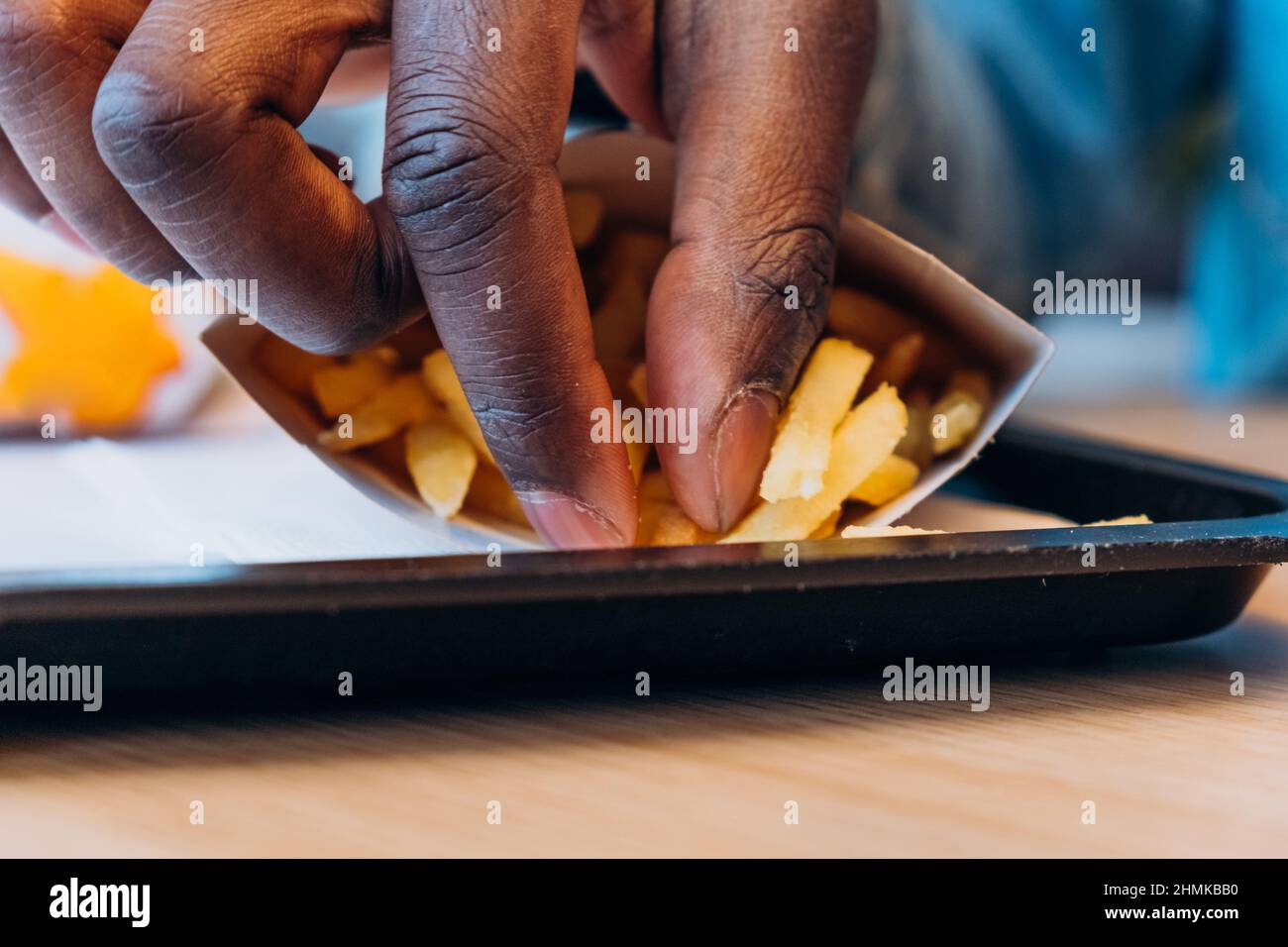 Ein junger afroamerikanischer Mann nimmt leckere frische pommes aus dem Papierbehälter an den Tisch im modernen Café, ganz aus der Nähe Stockfoto