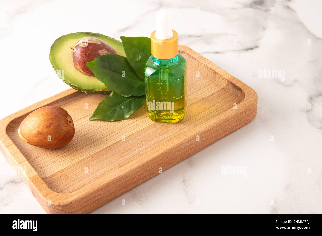 Avocado-Extrakt in einer farbigen Flasche auf einem Holztablett mit reifer Avocado. Feuchtigkeitsspendend, Vitamin E. Marmorhintergrund Stockfoto