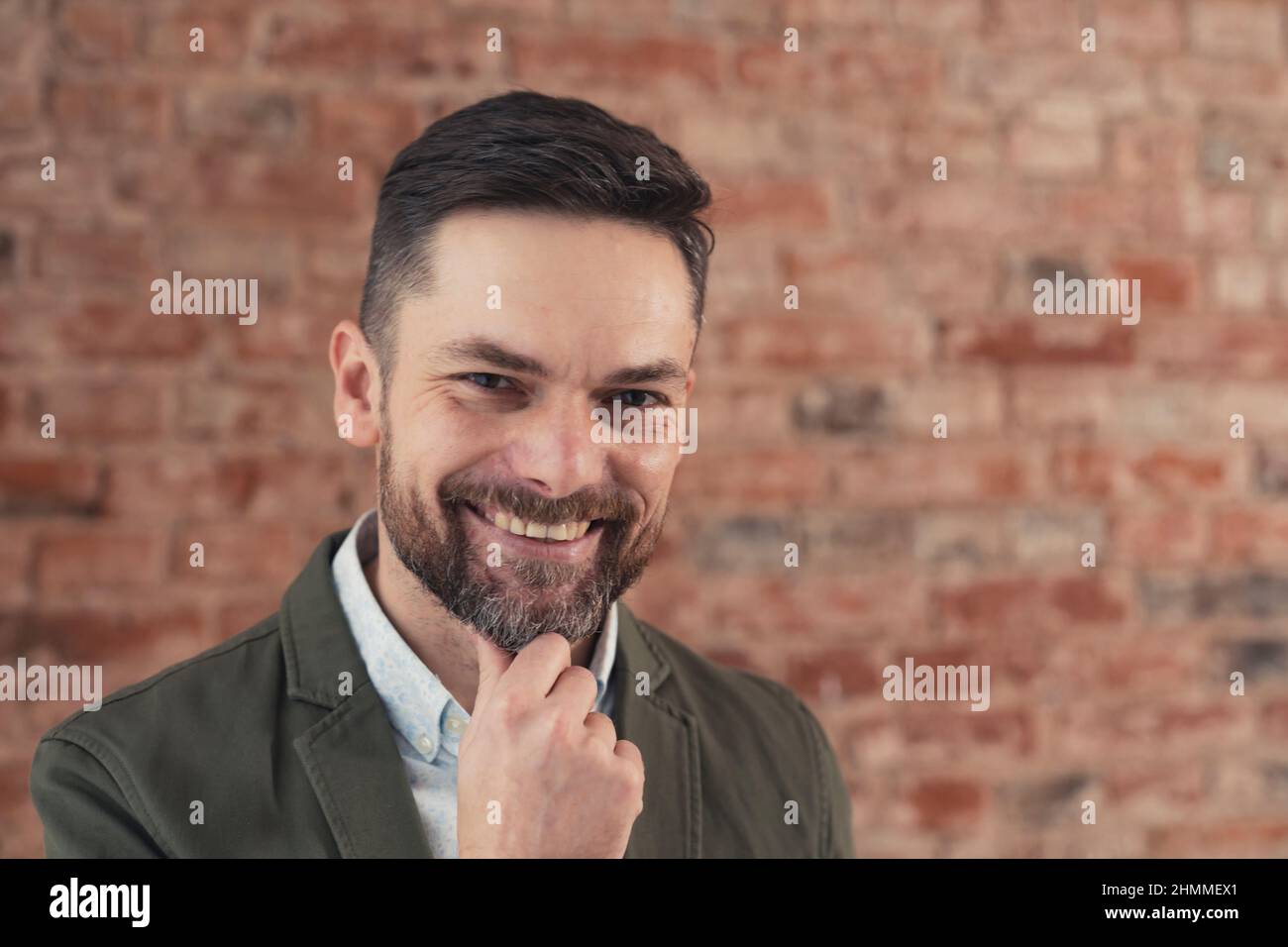 Kokett bärtiger kaukasischer Mann mittleren Alters grinsend und mit der Hand auf seinem Kinn vor Backstein auf die Kamera schauend. Hochwertige Fotos Stockfoto