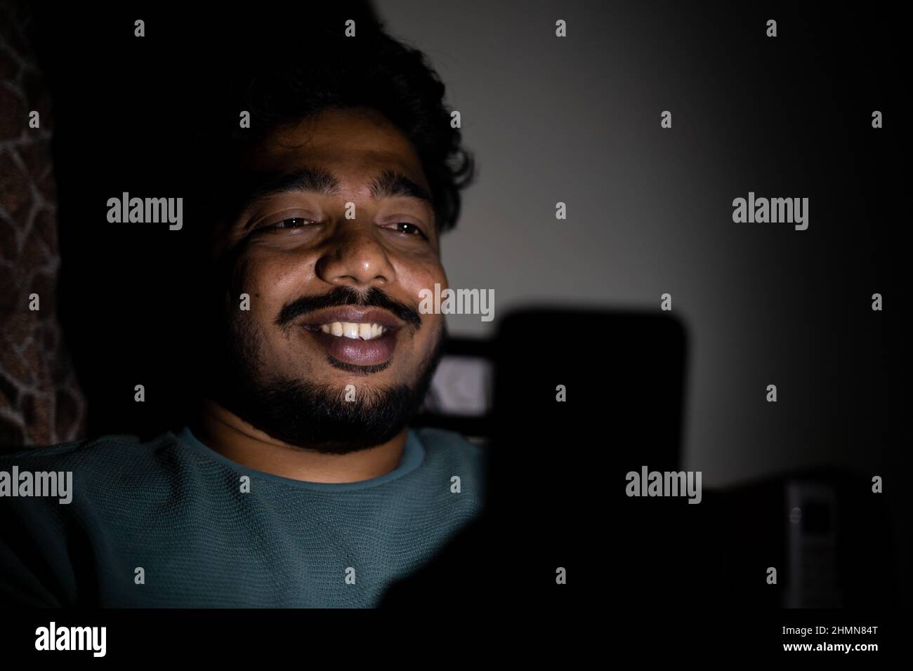 Glücklich lächelnder junger Mann, der in der späten Nacht mit den Augen reibt, auf dem Handy auf dem Bett chattet - Konzept des Online-Flirts oder -Dating und der sozialen Medien Stockfoto