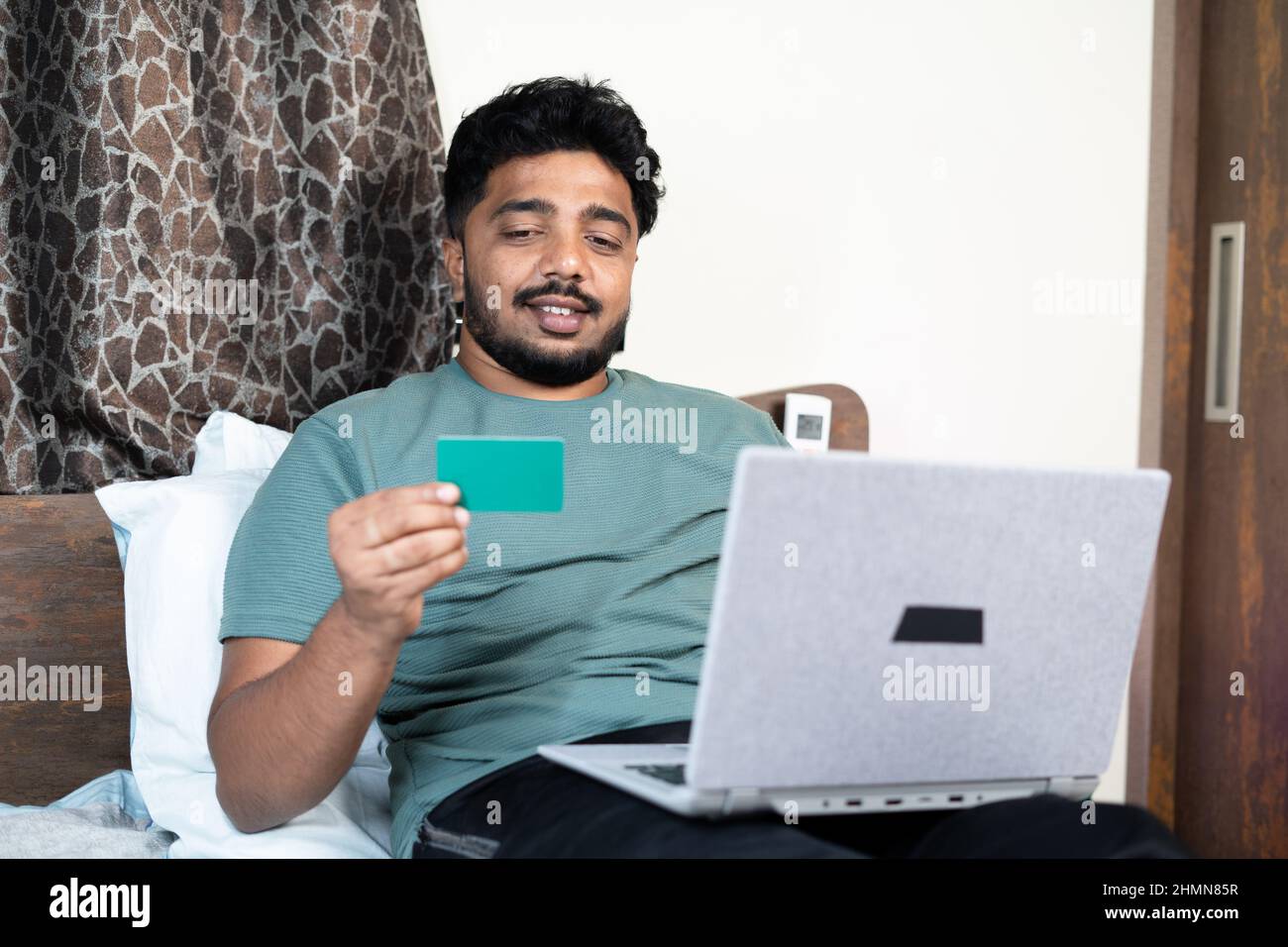 Junger Mann beschäftigt mit Bestellung oder Kauf mit Kreditkarte auf Laptop, während im Bett schlafen - Konzept der Online-Rechnungszahlung, E-Commerce-Shopping und Stockfoto