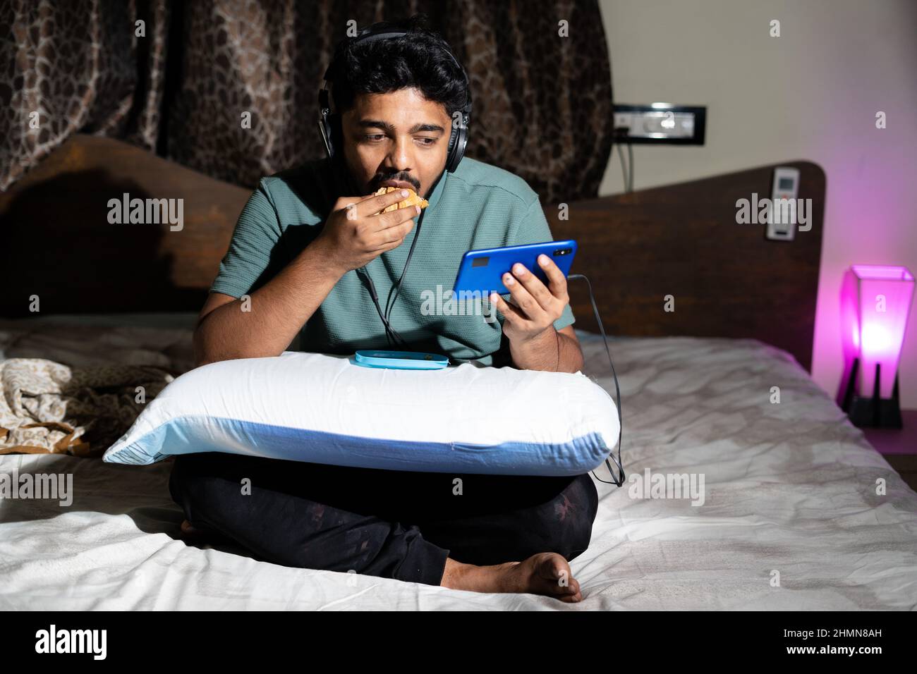 Junger Mann mit Kopfhörern, der Musikvideos auf dem Mobiltelefon genießt, indem er nachts Snacks isst – Konzept eines ungesunden Lebensstils, Online-Webserien gucken Stockfoto