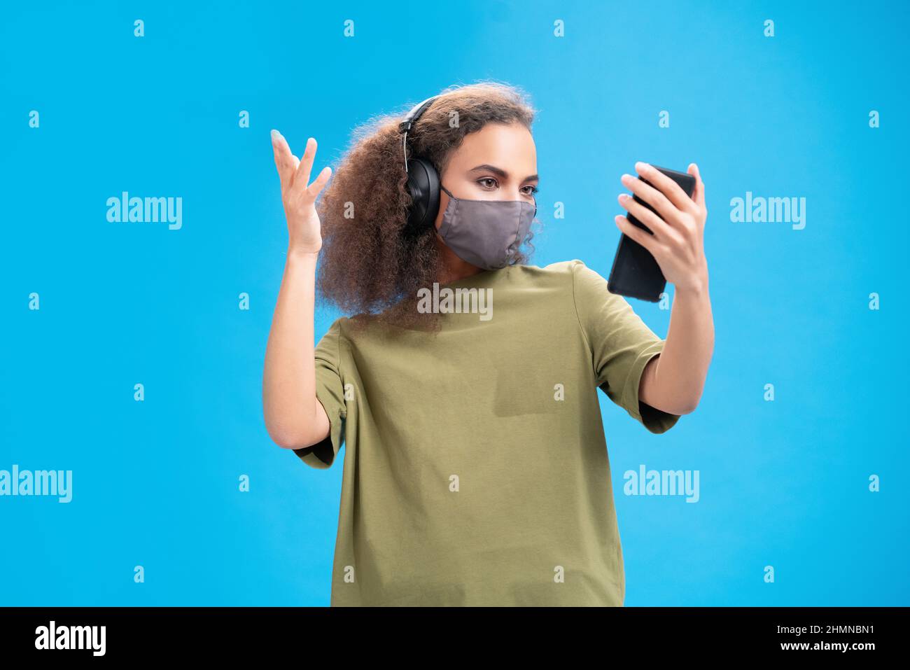 Junge afroamerikanische Mädchen in Schutzmaske Verwenden Sie Smartphone-Kamera, um mit Gebärdensprache oder Fingersprache mit Kopfhörern zu kommunizieren, isoliert auf blauem Hintergrund. Stockfoto