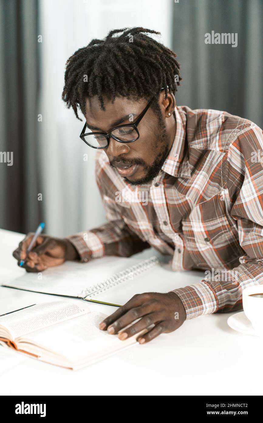 Afroamerikanischer Student liest Buch und macht sich Notizen in seinem Copybook, indem er seine Hausaufgaben macht. Portrait junger Mann in der Brille zu Hause oder in der Bibliothek studieren. Bildungskonzept. Stockfoto