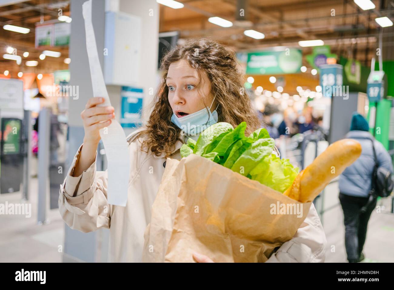 Eine junge Frau in einer medizinischen Maske sieht schockiert über einen Papierscheck in einem Supermarkt, der einen Papierbeutel mit Lebensmitteln, Preissteigerungen und Inflation hält Stockfoto