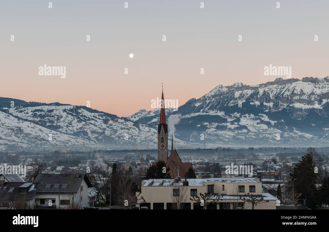 Schaan, Liechtenstein, 19. Januar 2022 Vollmondlandschaft in der Vormittagsszenerie hinter dem Turm einer katholischen Kirche in alpiner Landschaft Stockfoto