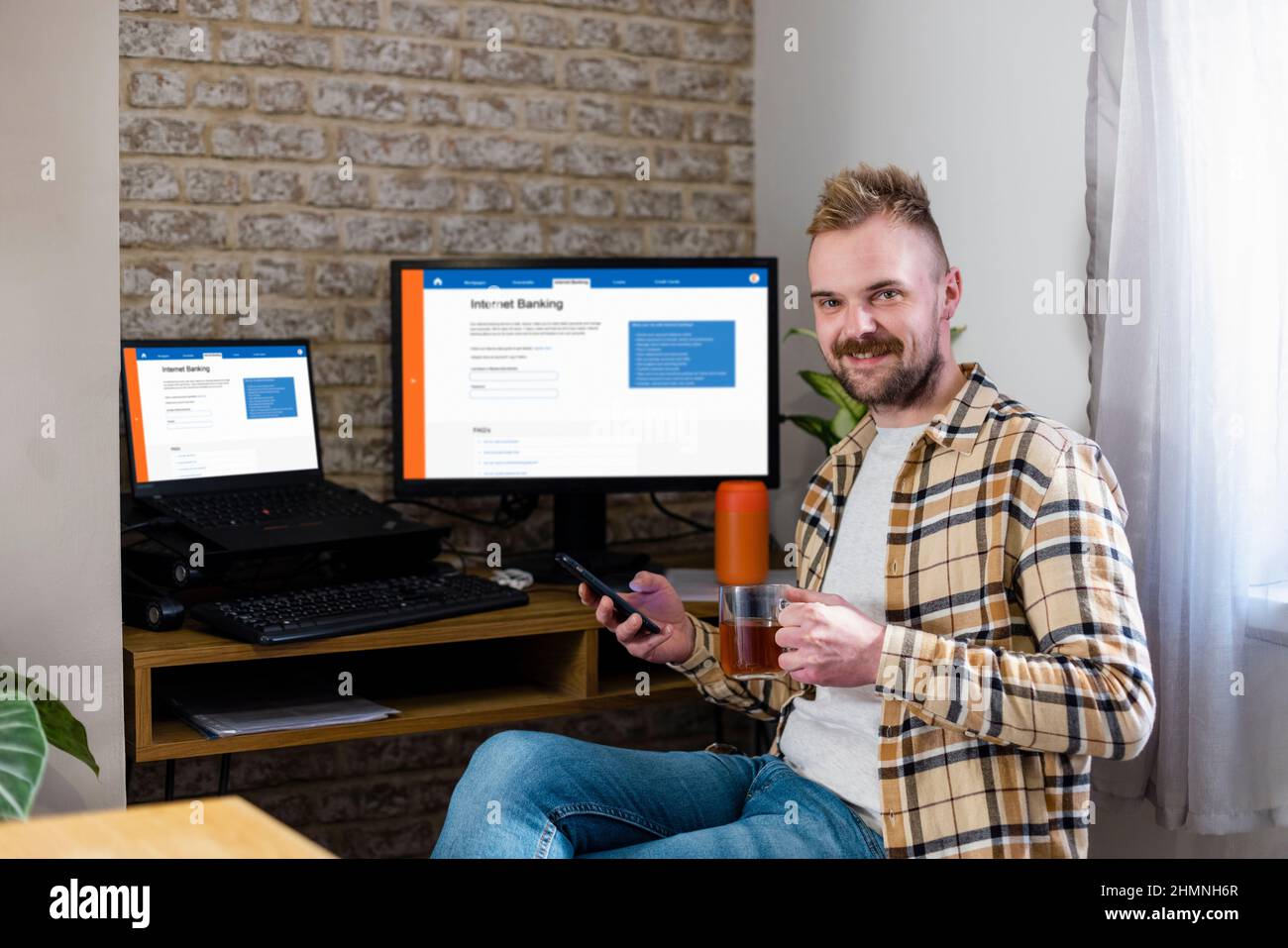 Ein Mann sitzt mit seinem Telefon an seinem Schreibtisch und blickt lächelnd auf die Kamera. Das Internet-Banking wird auf seinen Arbeitsbildschirmen angezeigt. Stockfoto