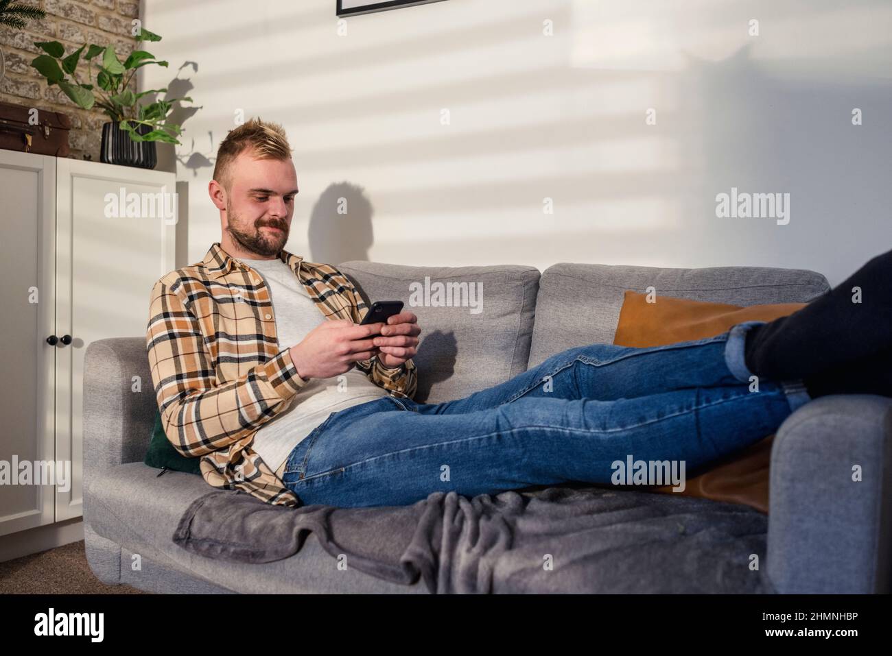 Eine Seitenansicht eines jungen Mannes, der entspannt auf seinem Sofa in seinem Wohnzimmer liegt und sein Smartphone benutzt. Stockfoto