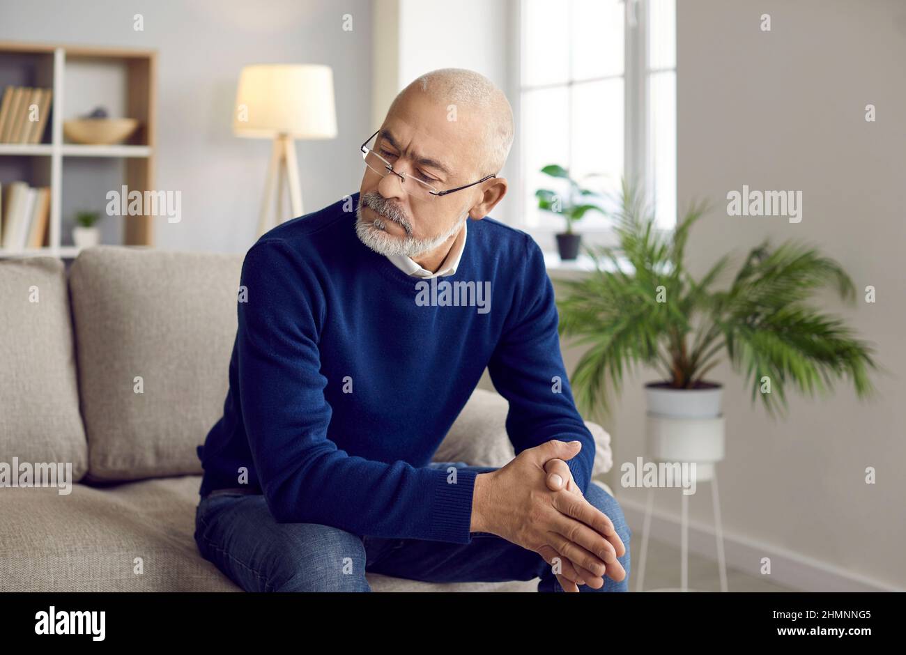 Älterer Mann in einer Brille sitzt zu Hause auf dem Sofa, blickt auf etwas und denkt nach Stockfoto