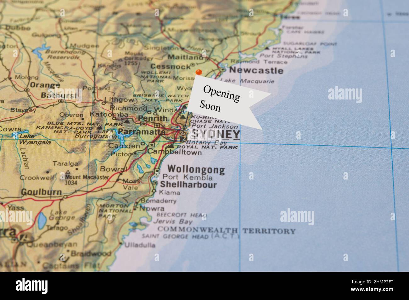 Eine Nahaufnahme einer kleinen Flagge mit dem Satz Opening Soon an einer Nadel, die in einer Illustration von Sydney Australia in einem Atlas platziert wurde Stockfoto