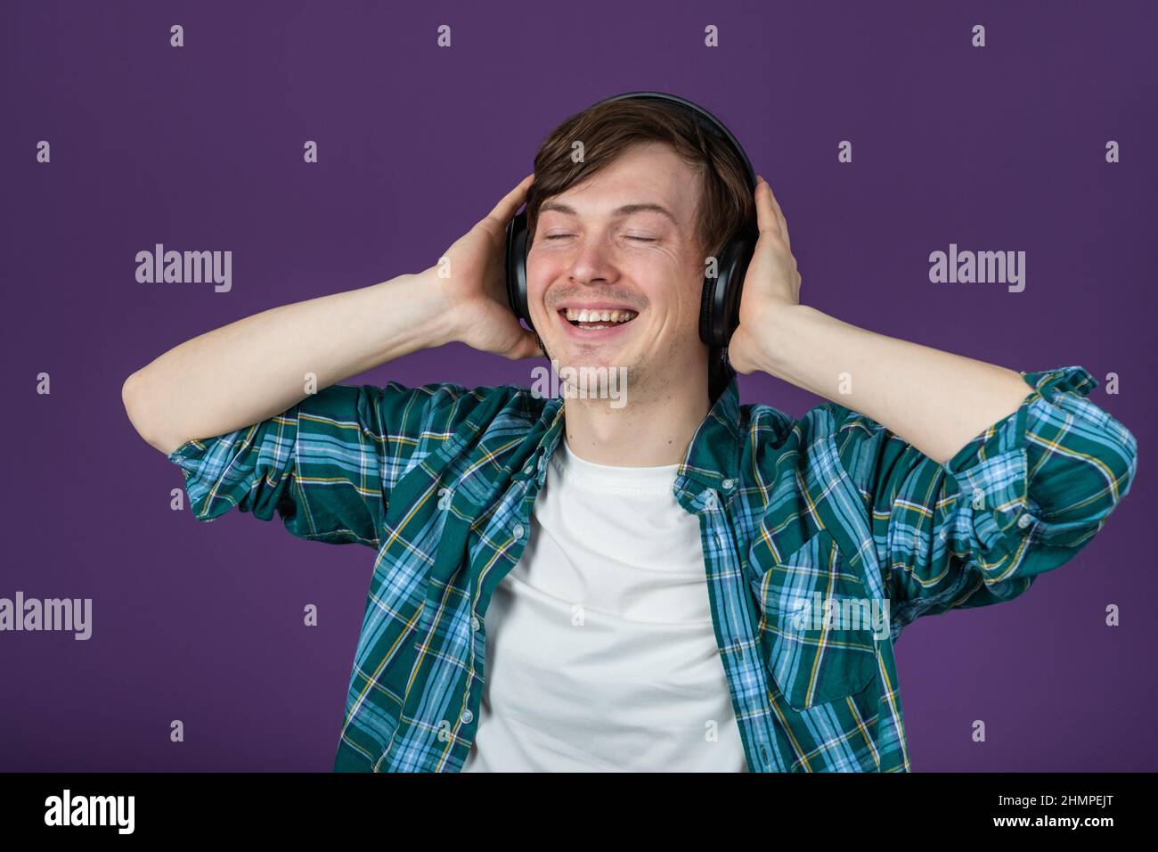 Ein glücklicher Mann in einem grünen Hemd hört Musik in Kopfhörern mit geschlossenen Augen und singt mit dem Kopf und den Händen auf einem violetten Hintergrund Stockfoto