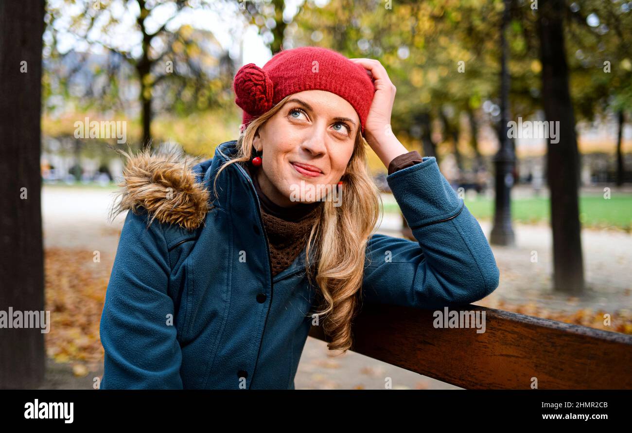 Eine junge kaukasische (deutsche) blonde Frau (Mädchen) mit blauen Augen  und einer roten Mütze und in ihren Gedanken, nachdenklich und leicht  lächelnd, sitzt auf einer Bank Stockfotografie - Alamy