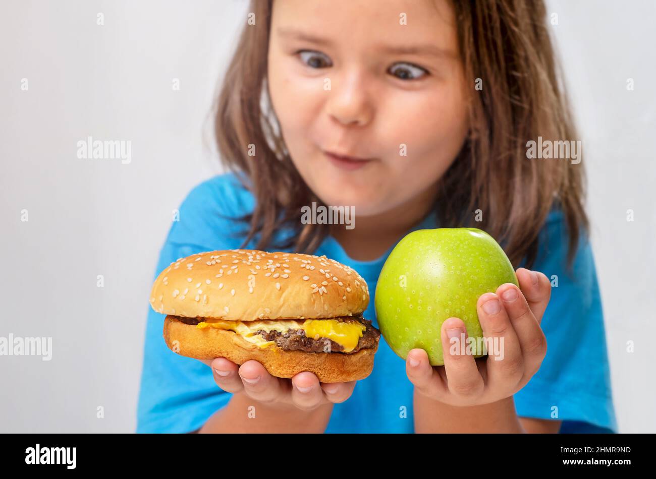 Das Mädchen schaut auf einen Burger, trifft die Wahl zwischen einem Cheeseburger und einem Apfel Stockfoto