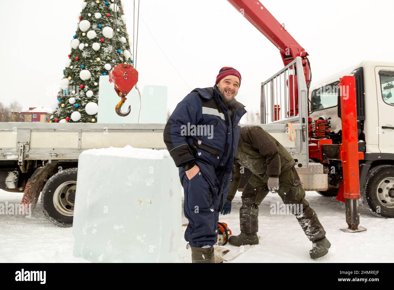 Porträt eines Arbeiters in blauer Jacke mit Kapuze am Haken eines LKW-Krans vor dem Hintergrund des Weihnachtsbaums Stockfoto