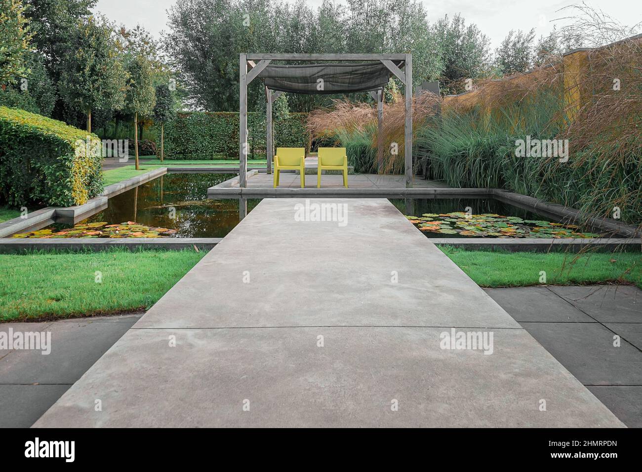 Zwei gelbe Stühle unter der Pergola, die von einem Pool aus Wasser umgeben ist, um die Sonne in einer Ecke eines schönen Gartens zu genießen. Stockfoto