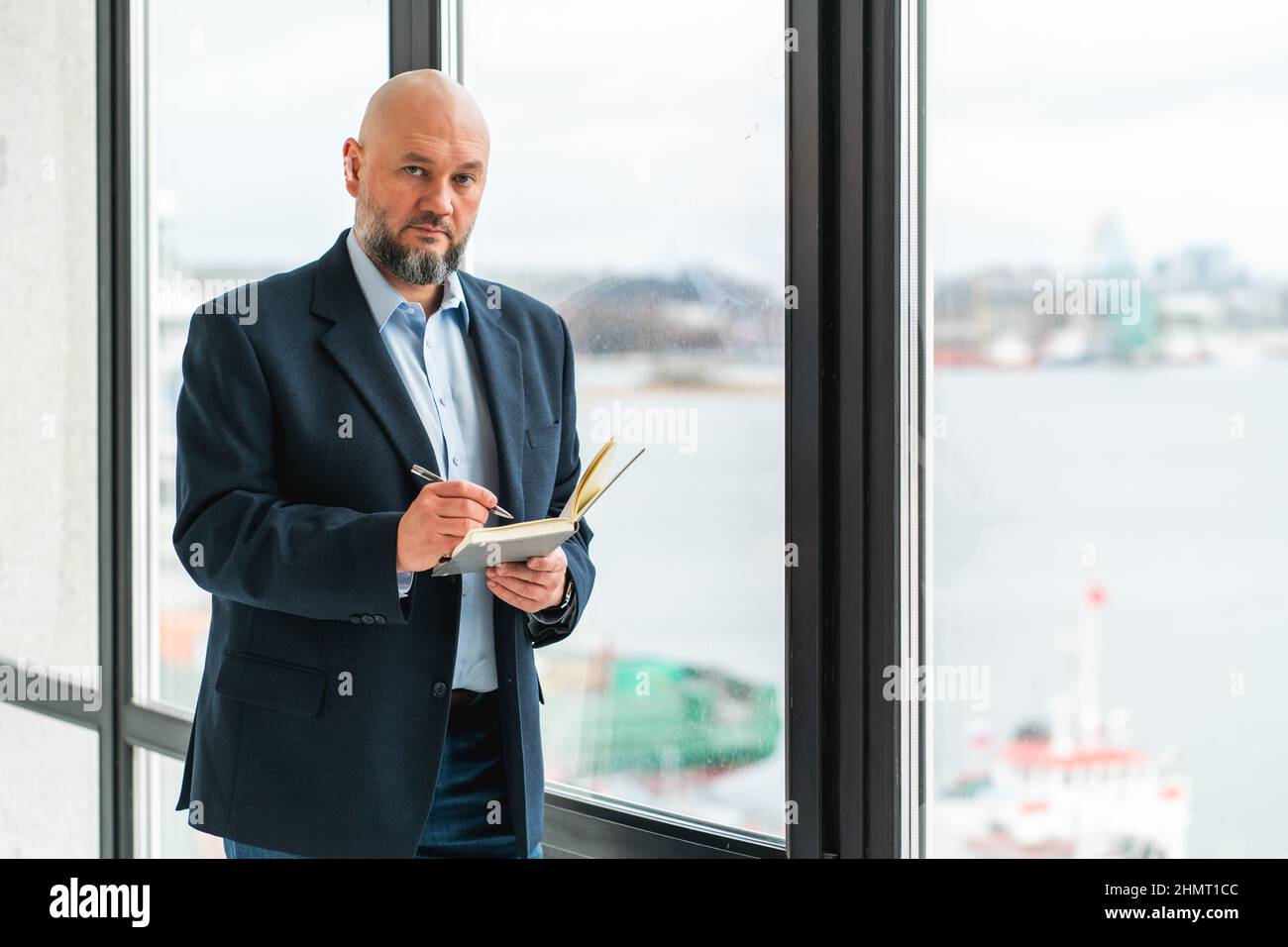 Seriöser, reifer Geschäftsmann im Anzug, der neben einem Panoramafenster mit Notebook steht Stockfoto