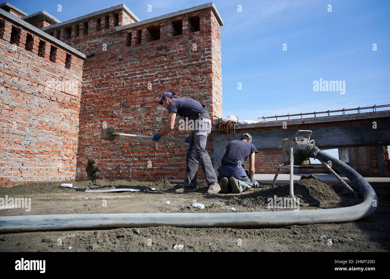 Zwei Arbeiter, die Zementgemisch, das von einer mobilen Mörtelpumpe bereitgestellt wird, auf die Arbeitsfläche auftragen. Installation von halbtrockenem Estrich draußen unter offenem blauen Himmel. Stockfoto