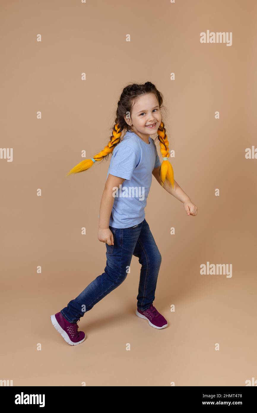 Junge unbeschwerte glücklich Mädchen mit gelben Kanekalon Zöpfe bewegen Arme und Beine, tanzen lächelnd Blick auf die Kamera auf beigem Hintergrund trägt blau t Stockfoto
