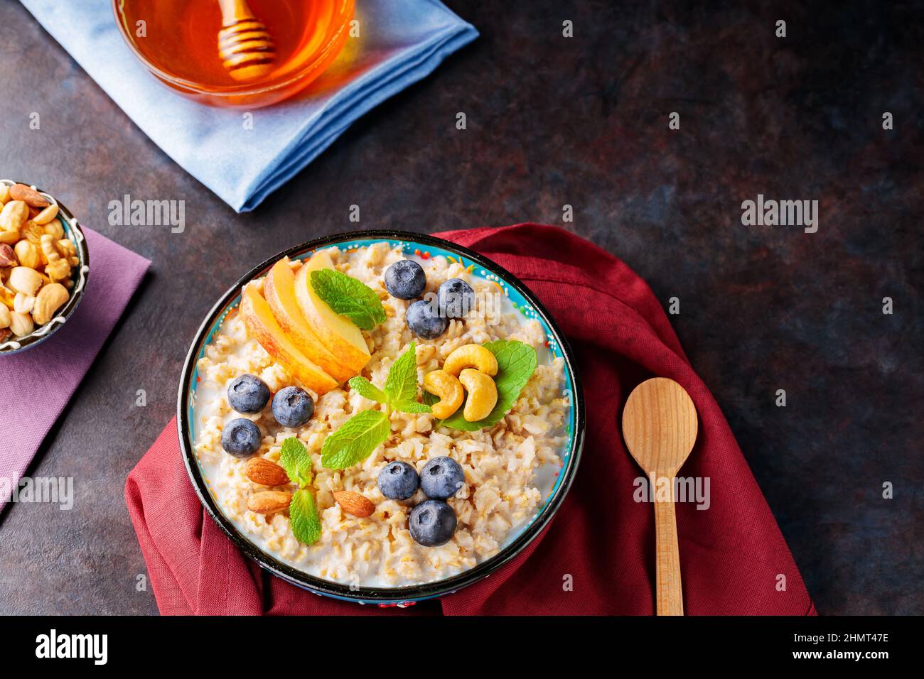 Haferbrei mit Joghurt, Beeren und Nüssen auf burgunderfarbener Serviette. Schüssel mit Haferflocken, Blaubeeren, Äpfeln und Honig auf dunklem Grund. Gesunde Brea Stockfoto
