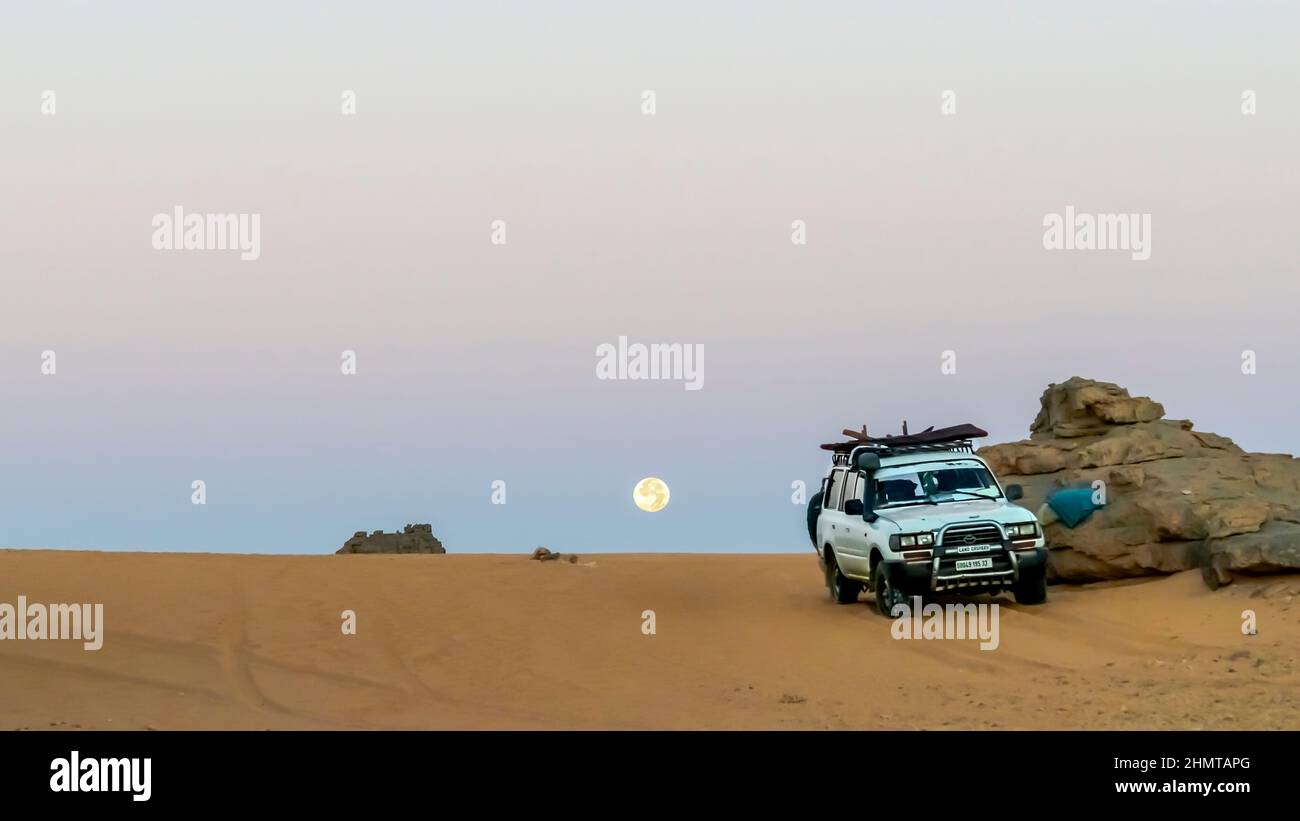 Der alte Toyota Landcruiser parkte an einem Felsberg in der Sahara mit Sand im Vordergrund. Farbenfrohe Morgendämmerung mit einem Vollmond, der im Hintergrund aufgeht. Stockfoto