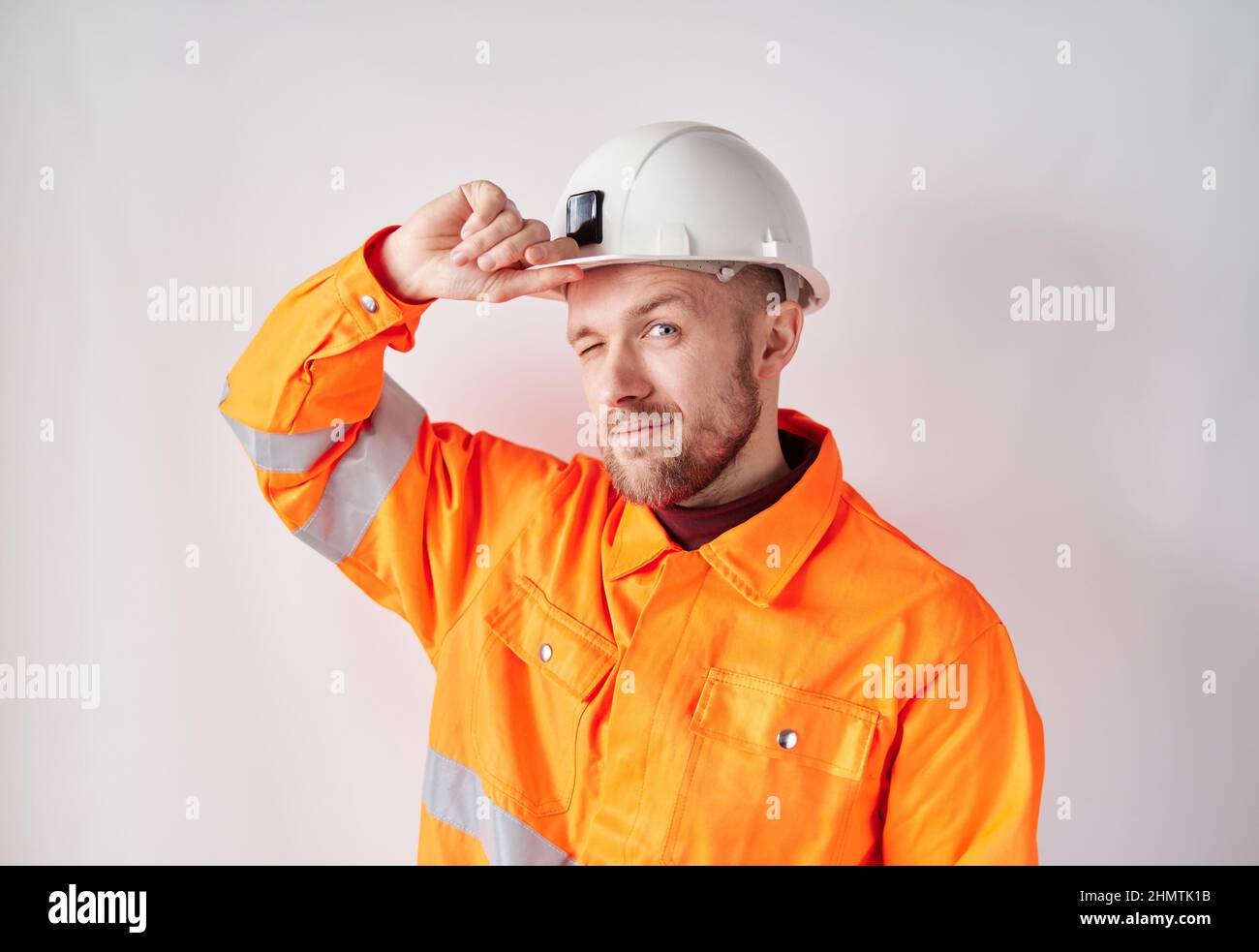 Fröhlich schauend auf Kamera junger kaukasischer Ingenieur, Arbeiter, Supervisor oder Bauunternehmer in weißem Helm und orangefarbener Jacke, der seinen Hardhut korrigierte. Konzept der Bauindustrie. Hochwertige Bilder Stockfoto
