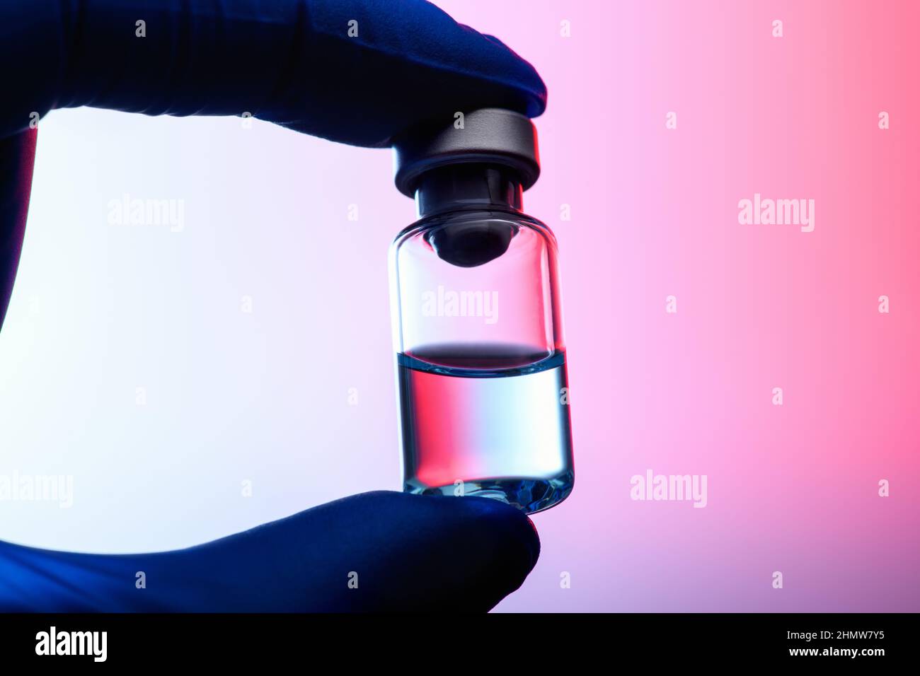 Der Coronavirus-Impfstoff in der Hand in einem medizinischen Handschuh mit rosa Hintergrundbeleuchtung, Silhouette. Impffläschchen gegen Coronavirus auf einem hellrosa Gradienten zurück Stockfoto