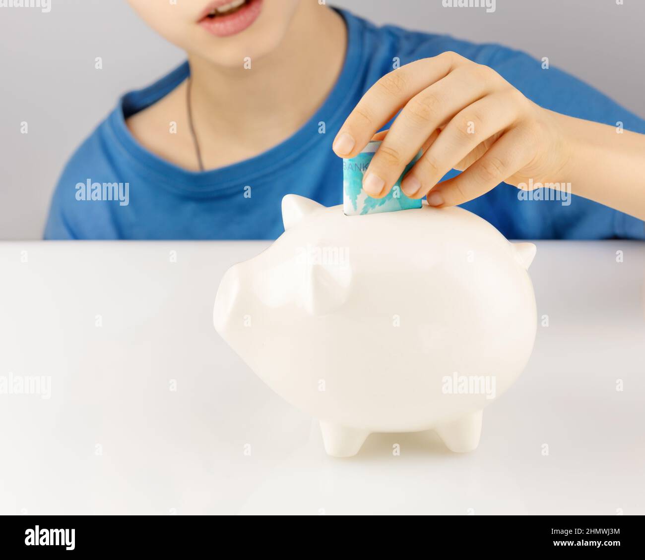 Das Konzept der Einsparung von Geld Investitionskonzept. Der Junge steckt vorsichtig Papiergeld in das weiße Sparschwein. Weichfokus Stockfoto