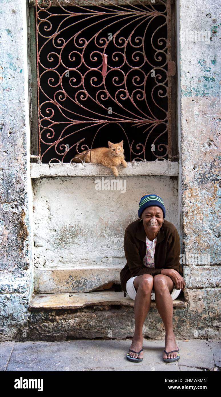 Die kubanische Frau sitzt auf der vorderen Stufe ihres Hauses mit ihrer orangefarbenen gestromten Katze. Stockfoto