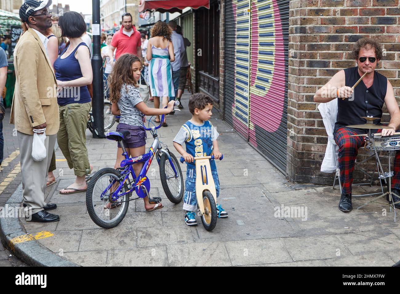 London, Großbritannien - 17. Juli 2020, Kinder, Geschwister auf Fahrrädern, hielten an, um Straßenmusikern auf dem Columbia's Sunday Flower Market zuzuhören Stockfoto