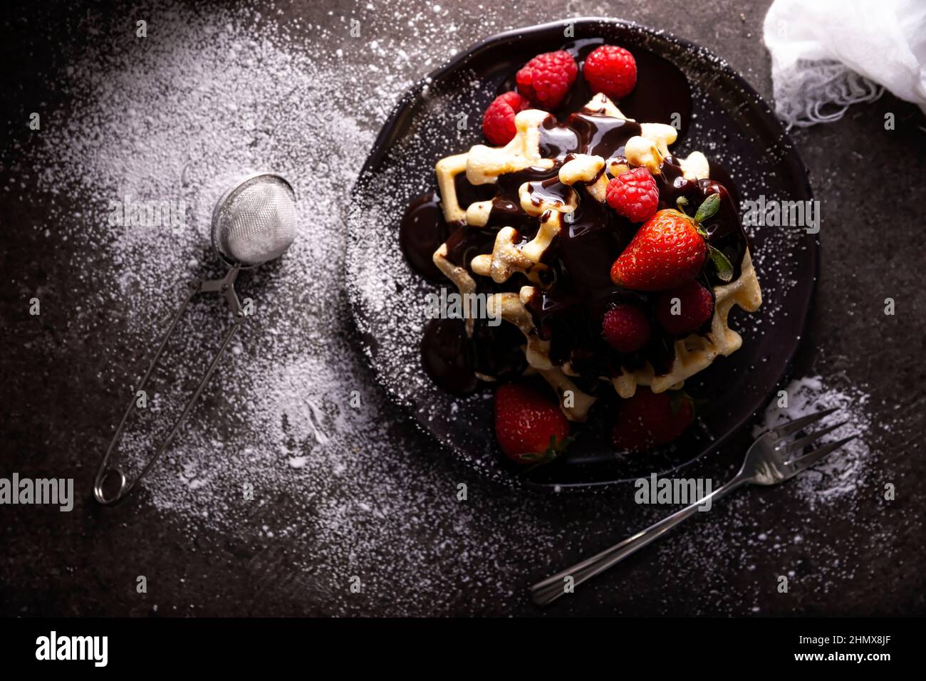 Blick von oben auf hausgemachte Waffeln mit Schokolade, Puderzucker, Erdbeeren und Himbeeren auf dunklem Gericht. Chaotisch aussehen flach liegen. Stockfoto
