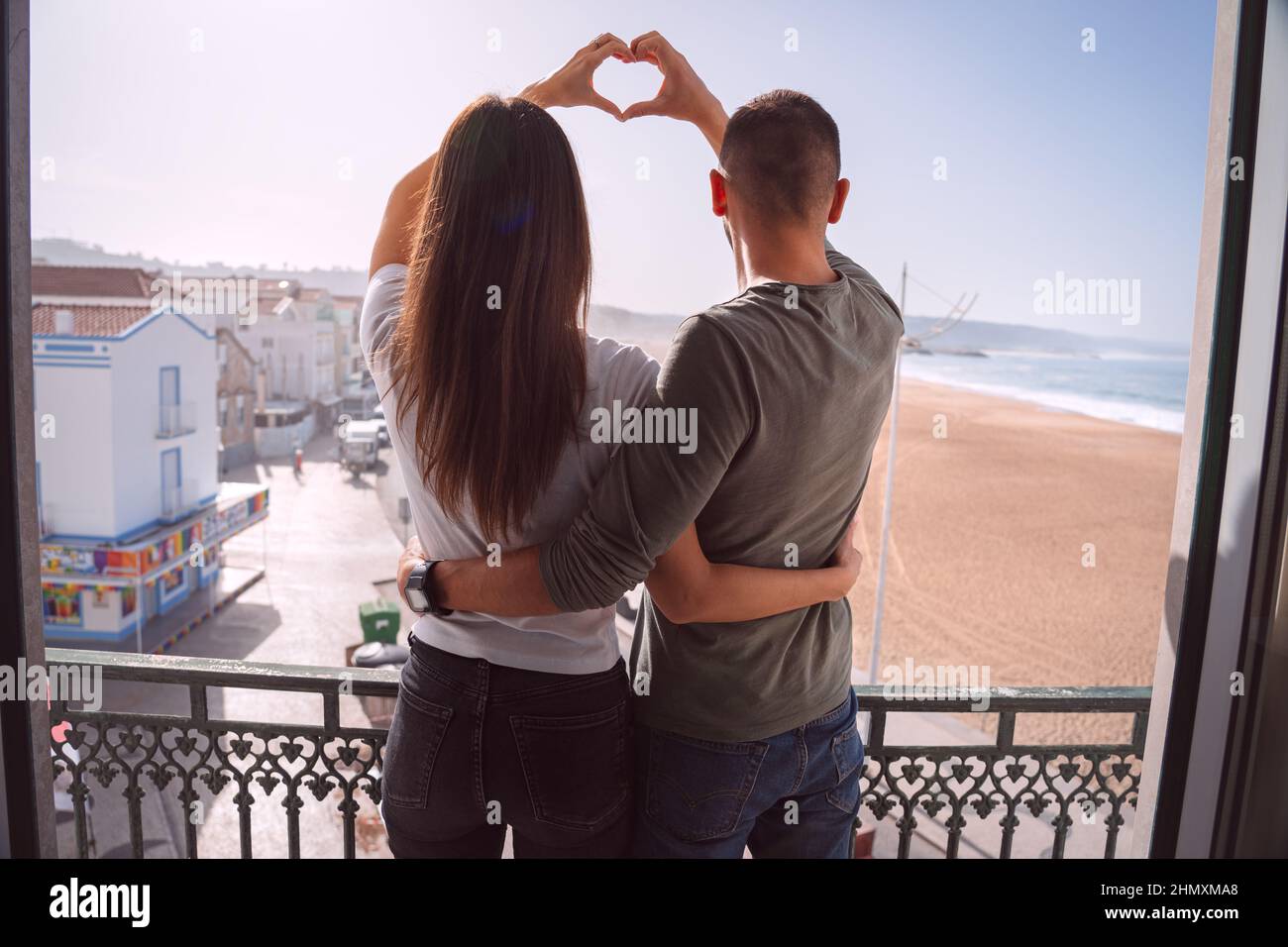 Junges Paar, das auf dem Balkon steht und mit den Händen Herzform formt. Verliebte Paare umarmen und verbringen den valentinstag zusammen Stockfoto