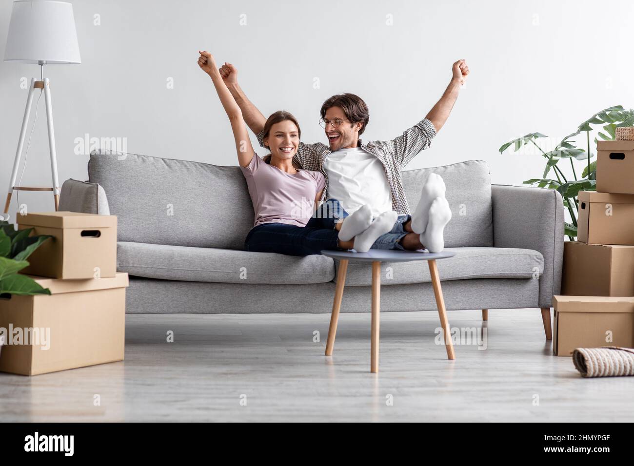Zufriedene kaukasische Millennials-Paare sitzen auf dem Sofa, ruhen sich aus, heben ihre Hände hoch und feiern den Kauf einer neuen Wohnung Stockfoto