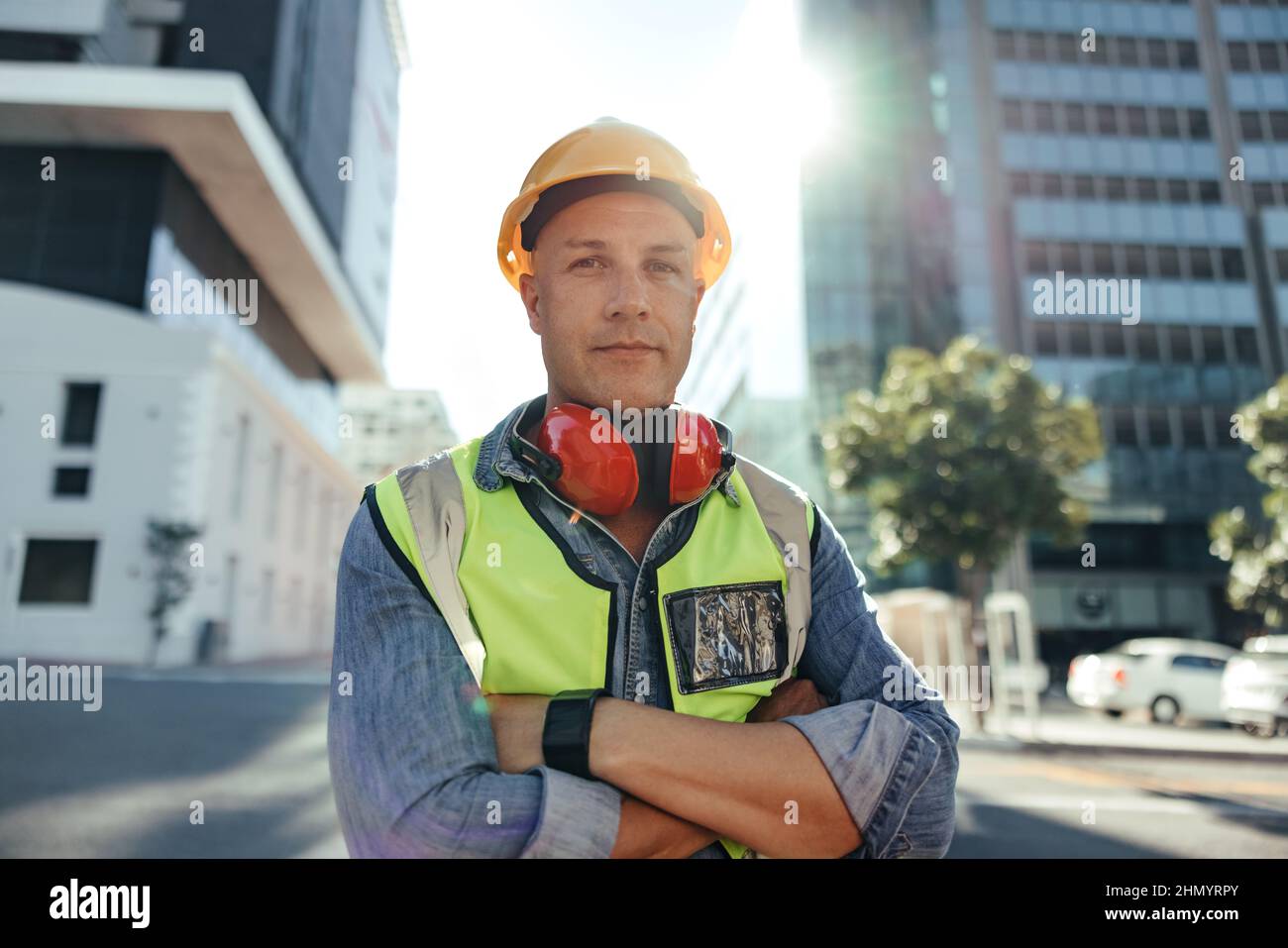Selbstbewusster Bauarbeiter, der die Kamera im Freien betrachtet. Der halbjährige Bauingenieur steht mit gekreuzten Armen in der Stadt. Arbeiter mit blauem Kragen Stockfoto
