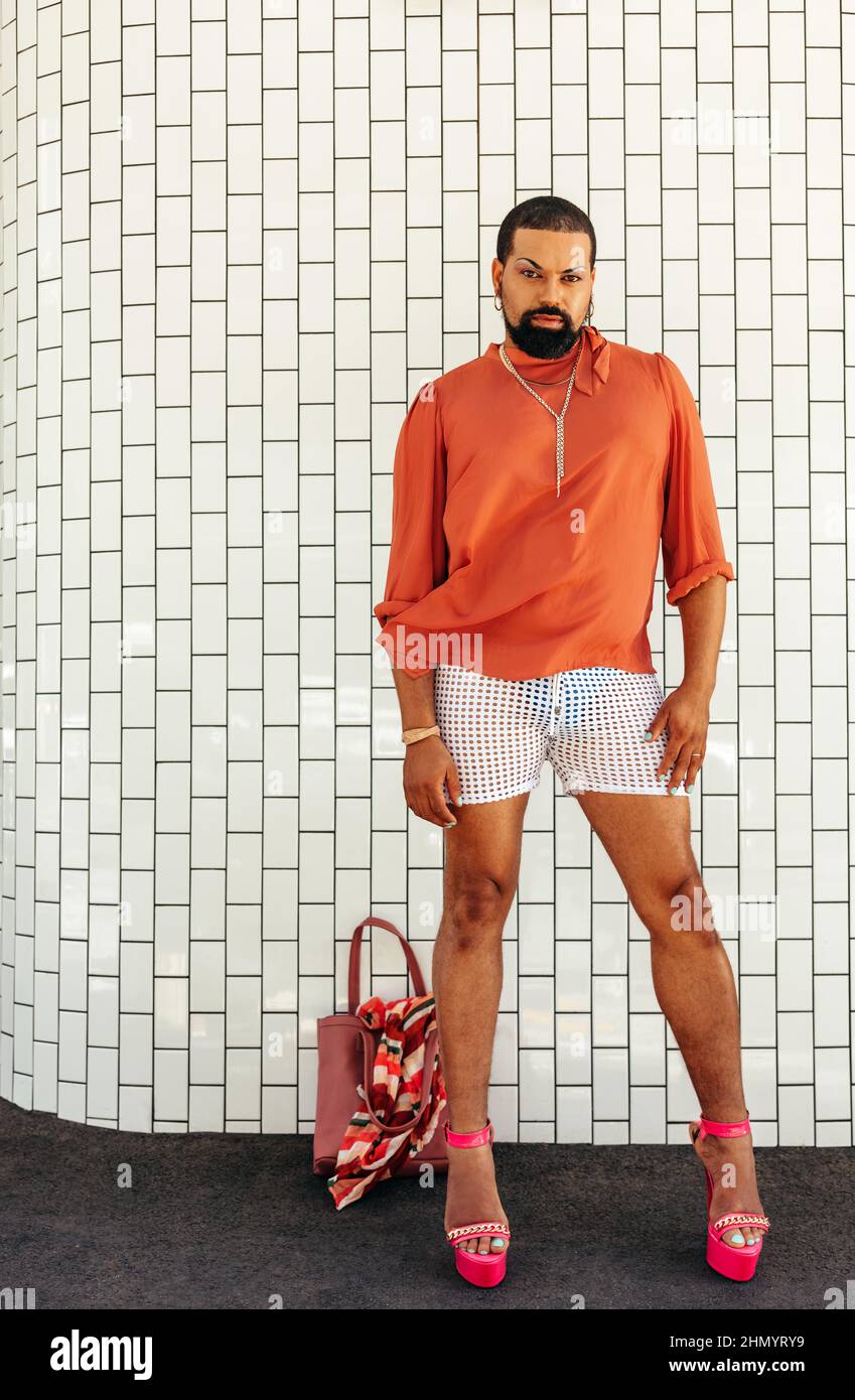 Elegante Drag Queen, die die Kamera anschaut, während sie vor einem weißen Hintergrund steht. Nicht konforme junge Mann trägt Make-up und Schmuck. Junger Mann Stockfoto