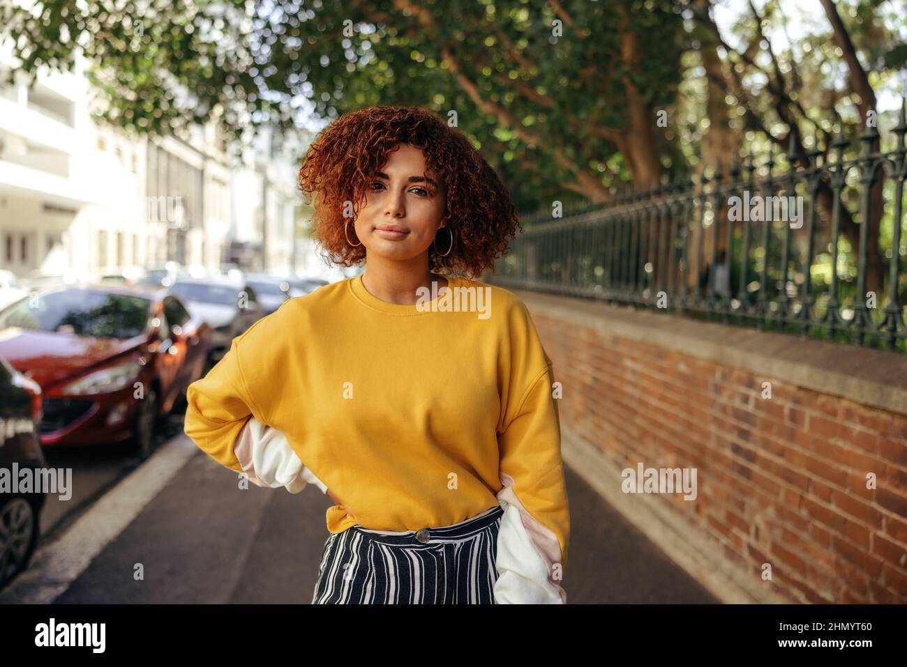 Sorgloses Teenager-Mädchen, das auf einem städtischen Bürgersteig mit der Hand auf der Hüfte steht. Junge Frau mit roten Haaren und einem Senf-Sweatshirt in der Stadt Stockfoto