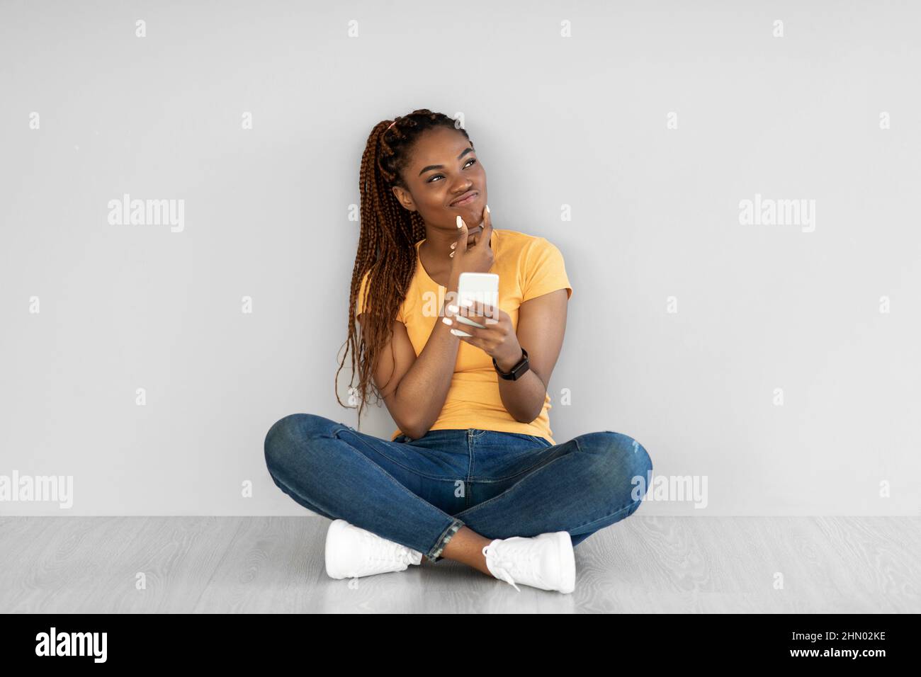 Nachdenkliche junge schwarze Frau, die mit gekreuzten Beinen auf dem Boden sitzt, Smartphone benutzt und über etwas gegen die graue Wand nachdenkt Stockfoto