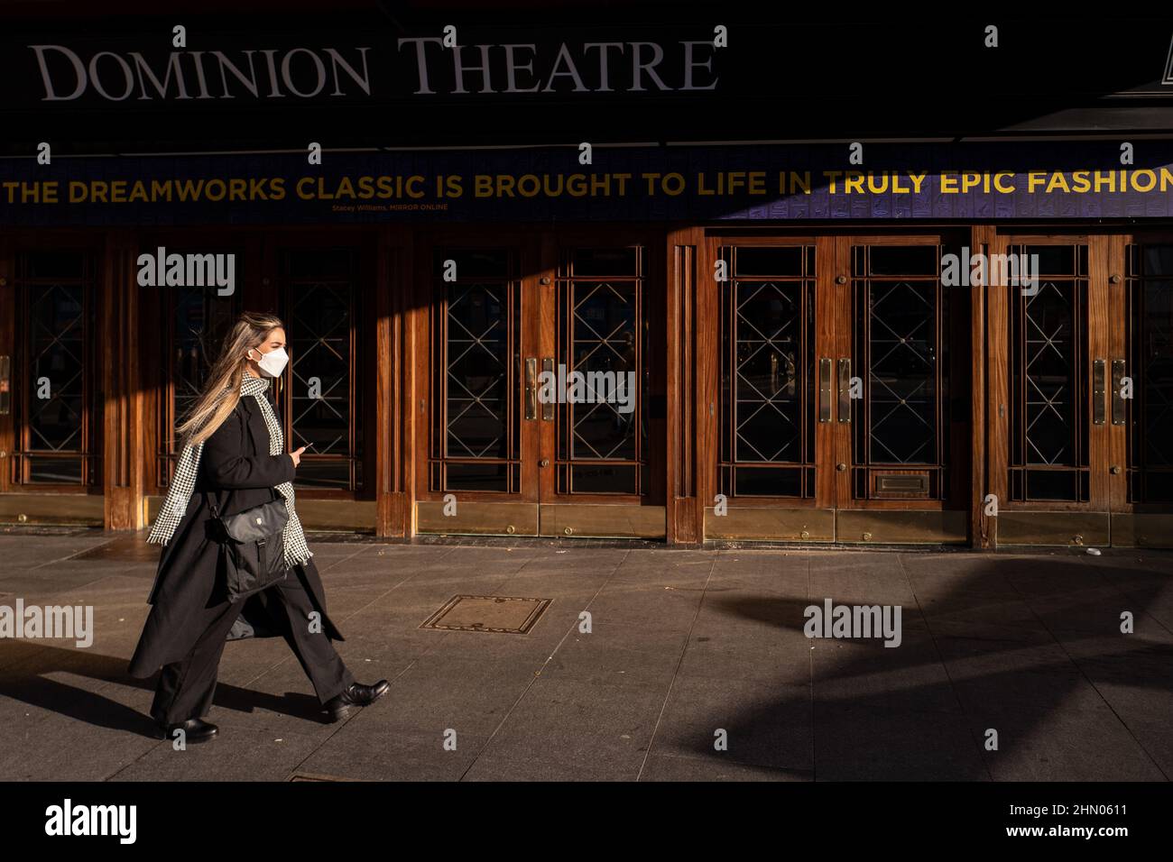 Eine Frau, die eine Gesichtsmaske, eine lange Jacke, einen Schal und eine Hose trägt, kommt weit vorbei am Londoner Dominion Theatre Stockfoto