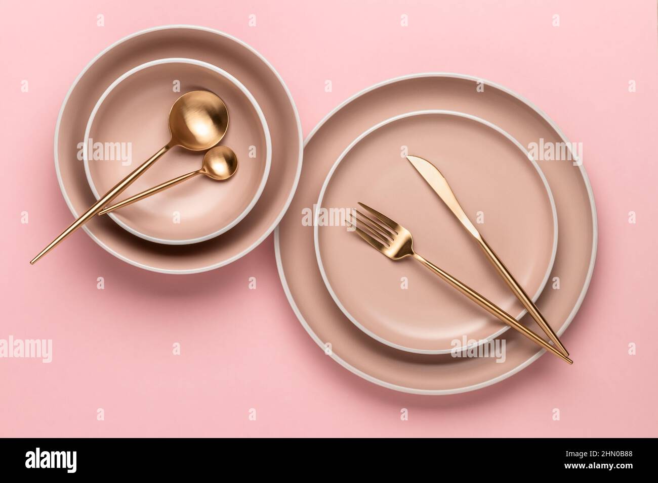 Geschirr und Utensilien zum Servieren und Essen von Mahlzeiten. Beigefarbene runde Teller mit Rand und goldenes Besteck auf rosa Hintergrund, Draufsicht. Moderne Keramik cr Stockfoto