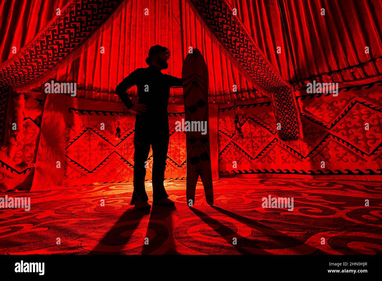 Silhouette von man Holding mit gespaltenem Schneeboard im Yurt Nomadenhaus gegen Teppich mit ethnischen Mustern, die von einem roten Licht im Inneren beleuchtet werden. Backcountry r Stockfoto