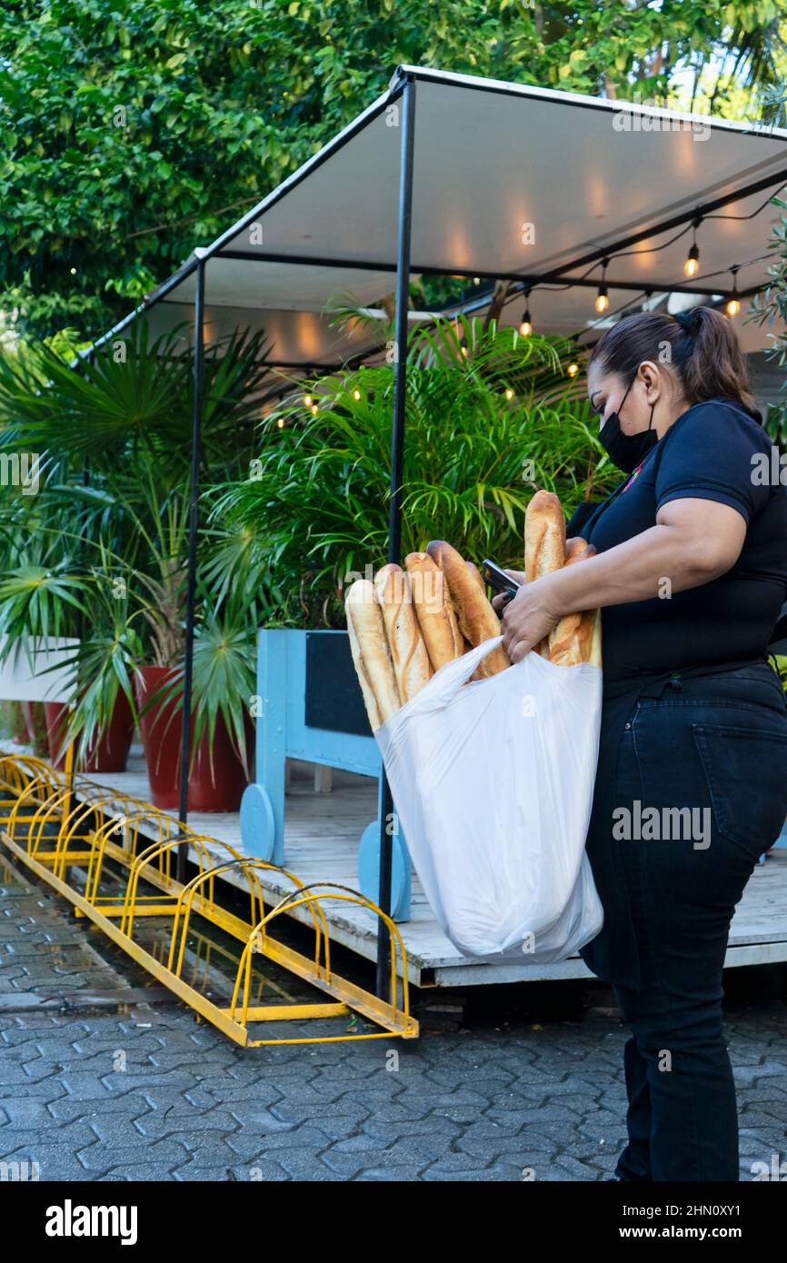 Die junge Mexikanerin mit einer Maske von covid19 und einer Tasche voller französischer Baguettes nutzt ihr Smartphone Stockfoto