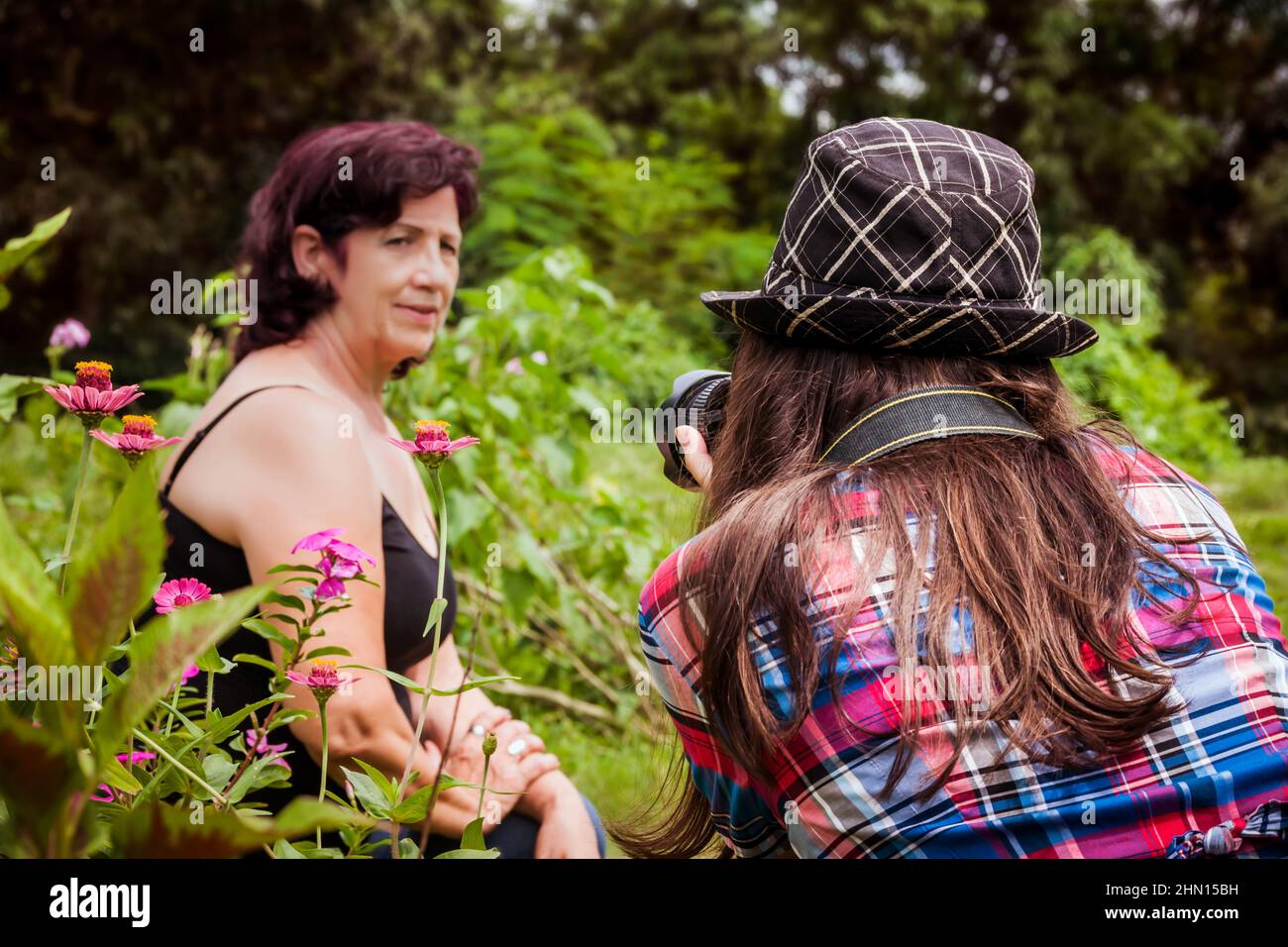 Nahaufnahme des Rückens einer jungen Fotografin mit langen braunen Haaren und einem Hut, der mit ihrer Linse auf eine Erwachsene lateinische Frau in einem Garten zielt Stockfoto