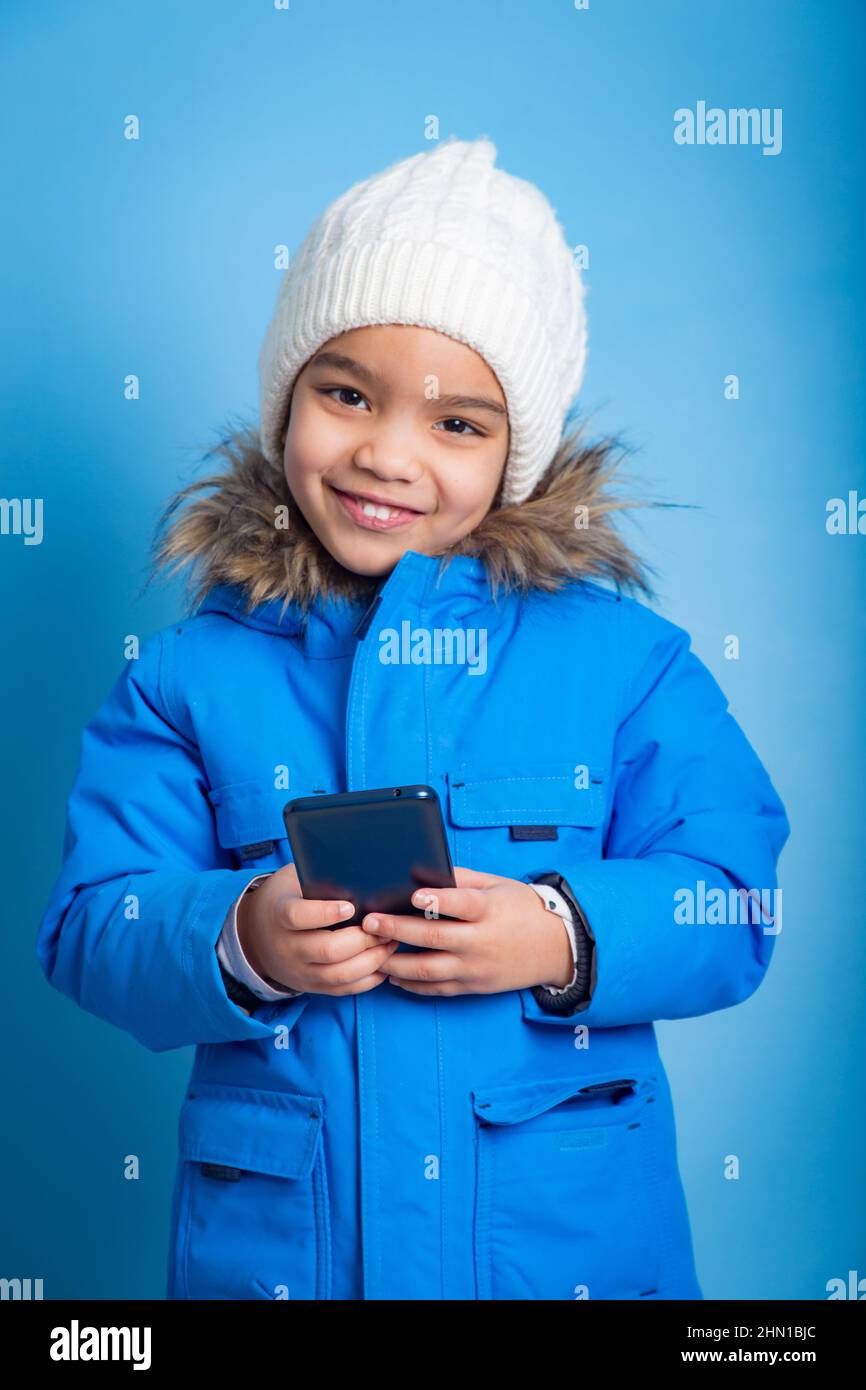 Kleines Mädchen mit einem Smartphone, das einen Winteranzug auf blauem Hintergrund trägt Stockfoto