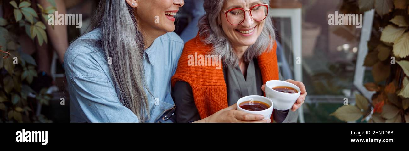 Lächelnde reife asiatische Dame mit Freund clink Tassen Kaffee sitzen in der Nähe des Cafés Stockfoto